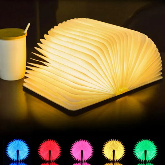 Foldable book-shaped lamp emitting a warm glow on a dark surface with a cup beside it, and color options displayed below.